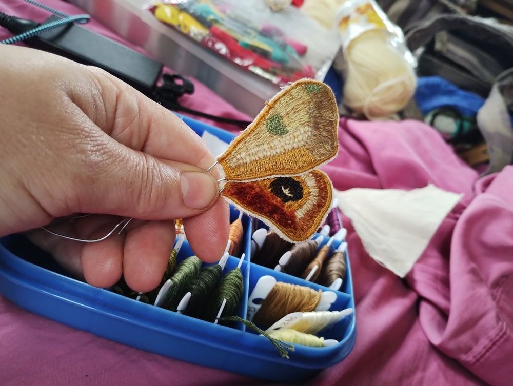 A hand holding a right-side pair of embroidered moth wings in tans and golds. The edges are still untrimmed. In the background is a bunch of embroidery floss.