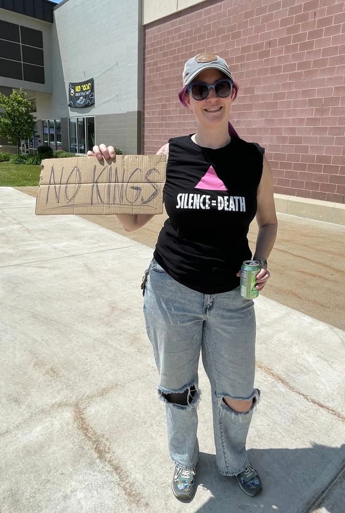 Jessi is holding up a No Kings sign written on cardboard. She's wearing torn jeans and a black ACT UP t-shirt that has a pink triangle and "Silence = Death" beneath it.