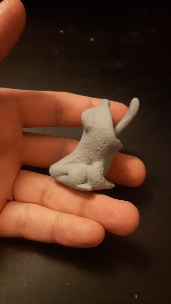 Frog figurine from the side. Head is pointing upwards, with mouth open and tongue extended. Proportionally tiny body and massive head. Stippled with dints over its skin for texture. Unpainted.