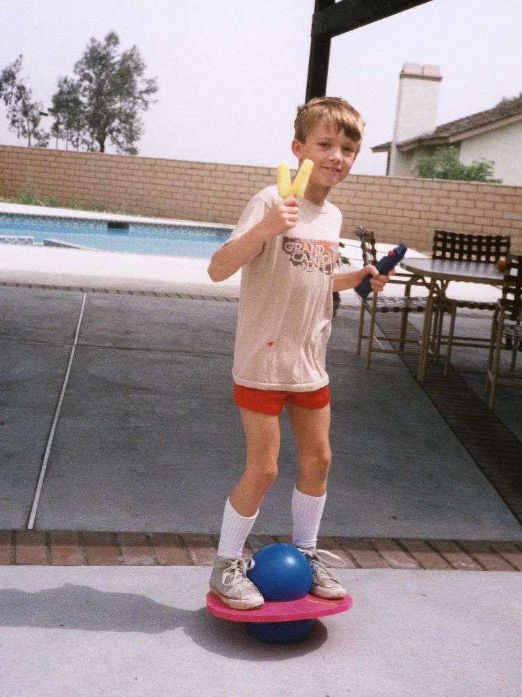 Young boy on a Pogo Ball holding a banana popsicle and a lightsaber hilt, wearing red short shorts with pulled-up white socks, and a Grand Canyon T-shirt.