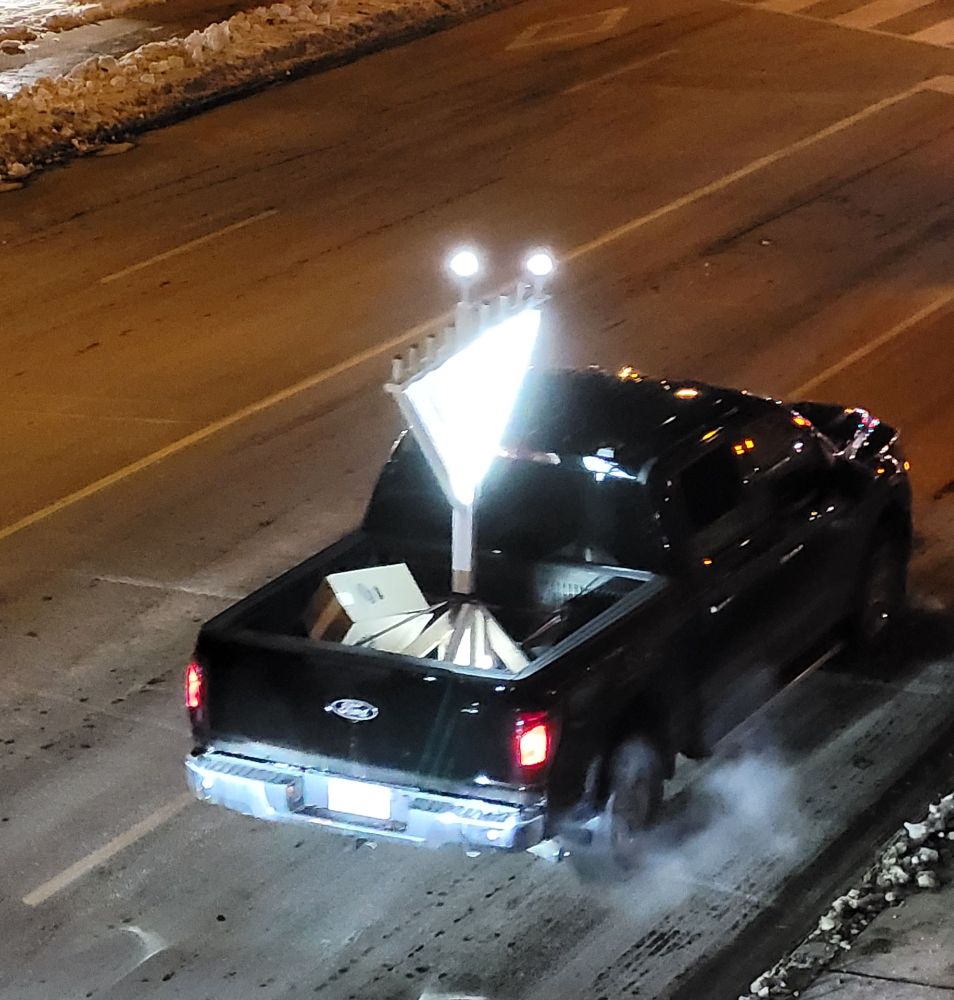 A pickup truck with a large light-up menorah in the back of its flatbed. A light on the outermost lamp is illuminated, as is a centre bulb representing the shamash.