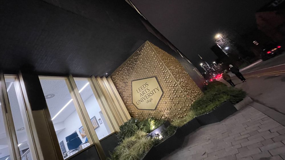 Outside the Leeds Arts University at night. People walking by. Lights of the city centre in the background, and a gold embossed university logo in the centre of the frame.