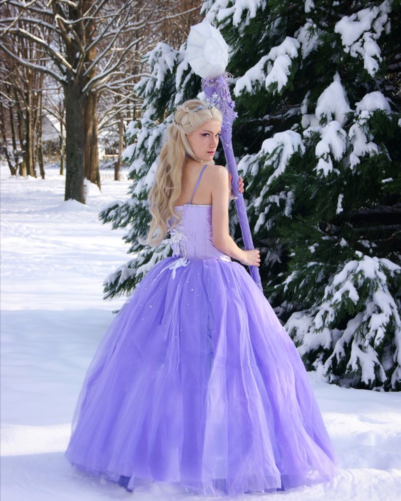 A woman stands in a snowy landscape. She is dressed in a purple, long, sleeveless gown. She stands with her back to the camera, has blonde hair and holds a huge purple wand with a white crystal on top.