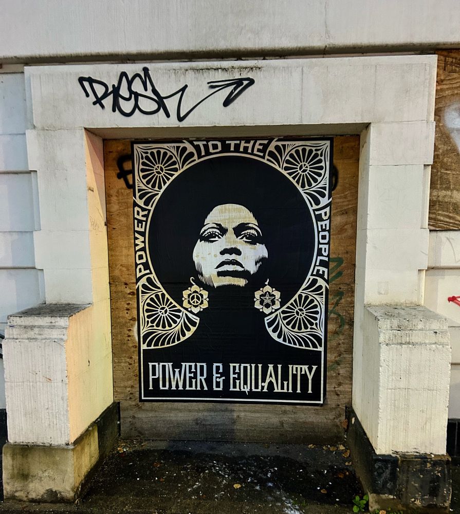 a large, black-and-white poster of a black woman with a powerful, contemplative expression. Surrounding her are intricate floral designs, adding to the bold visual impact. The words "POWER TO THE PEOPLE" and "POWER & EQUALITY" are displayed prominently in the artwork, emphasizing themes of social justice and empowerment. The piece is framed within a wooden board covering a doorway, suggesting it a somewhat makeshift setting. Above the mural, there’s a graffiti tag that reads "ROSE," adding an extra layer of street art culture to the scene.