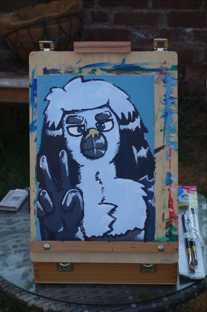 The same painting, sitting on an easel in a garden. It's fairly close up, so you can't see too much of the background, but you can see some brushes sitting next to it, some flower baskets behind, and grass and a brick wall.