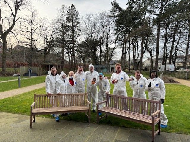 A group of Hilti volunteers, dressed in white protective suits, stand outdoors smiling and posing with two fully sanded wooden benches. They are in a green garden area with trees and a pathway in the background.