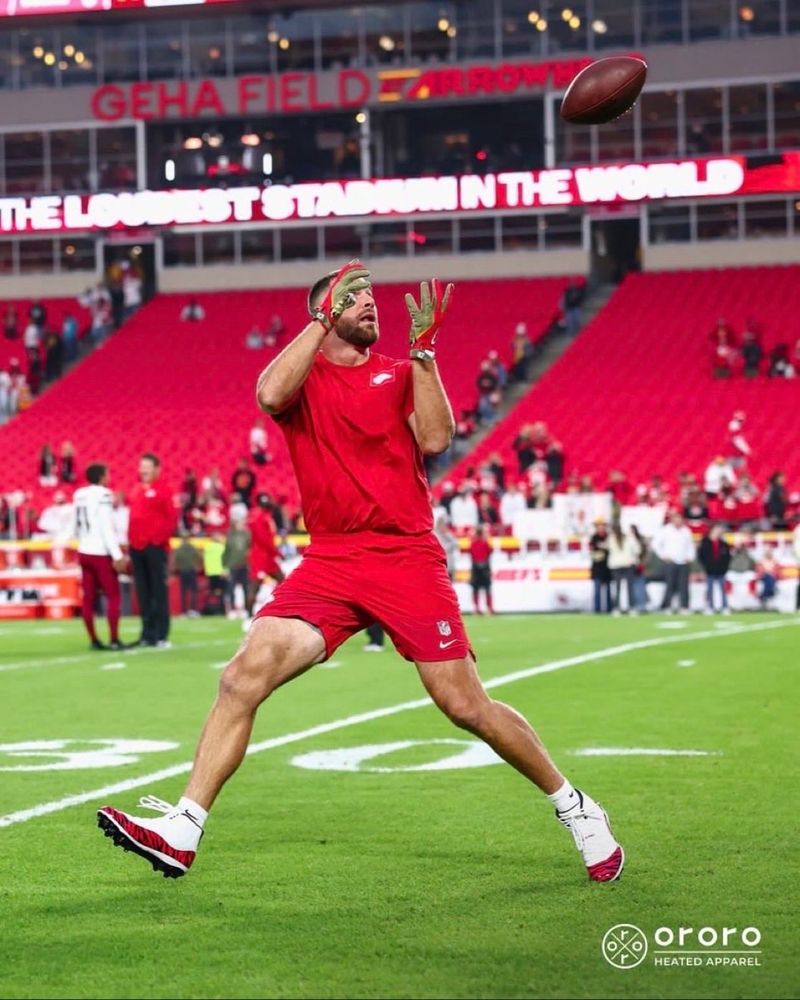 Travis Kelce in Chiefs red shirt and shorts. His is warming up on the field before the game. He is jumping to catch a football.