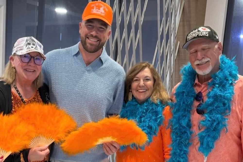 Donna Kelce holding an orange feather fan, Travis Kelce wearing a blue polo and orange hat and also holding an orange age feather fan. Two family members in orange shirts and teal boas.
