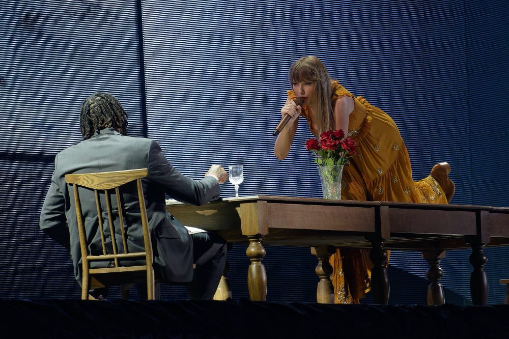 Photo from the tolerate it performance at the Eras Tour. Taylor is wearing a orangey yellow dress and is crawling across a large wooden table while singing to a man seated with his back to the camera.
