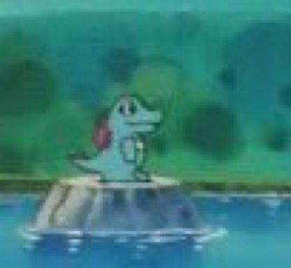 low res screenshot of a tiny Totodile in the Pokémon anime standing on a rock in a pond 