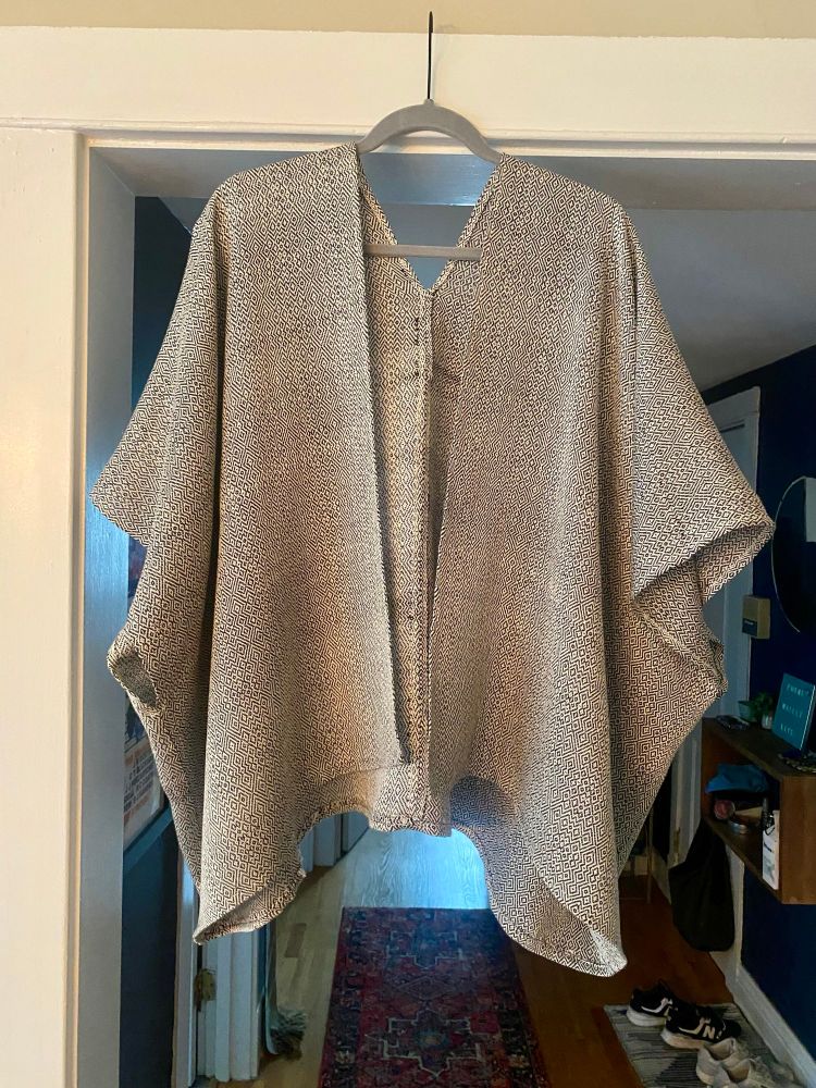 A loose, flowy, handmade top made from the black and white handwoven fabric. It is hanging from a wooden clothes hanger in a doorway. 