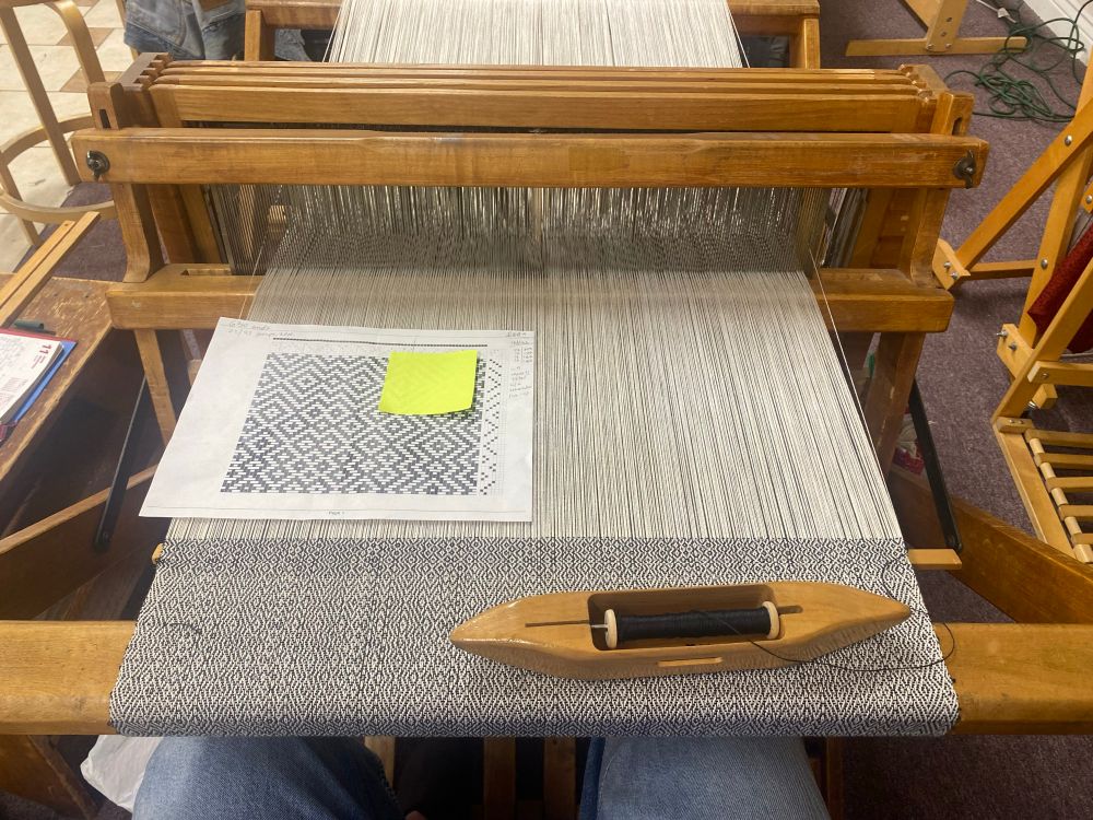 A woven work in progress on a wooden floor loom, in a twill pattern made from black and white yarn. A paper pattern sits on top of the work, and there is a wooden boat shuttle with a black bobbin. 