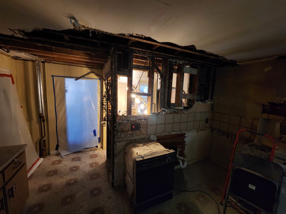 a kitchen wall torn down enough that you can see through it to the windows on the adjoining bathroom