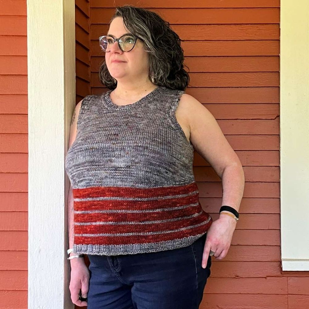 a white-skinned woman with dark hair and glasses is wearing a handknit tank top that's mostly gray with chunky reddish stripes on the lower half; she's standing against an orange wall, looking off to the side of the frame