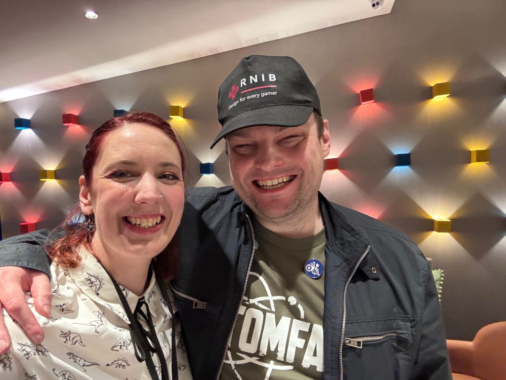 Searra shares a quick selfie ans hug with Sightless Kombat. Searra is on the left and wearing a white shirt with tiny dinosaurs printed on it and has red hair. Sightless is stood beside her to the right wearing a green Atomfall T-shirt, black jacket and a cap with the logo for RNIB on it.

Behind them is a wall covered in blue, yellow and red lights. 