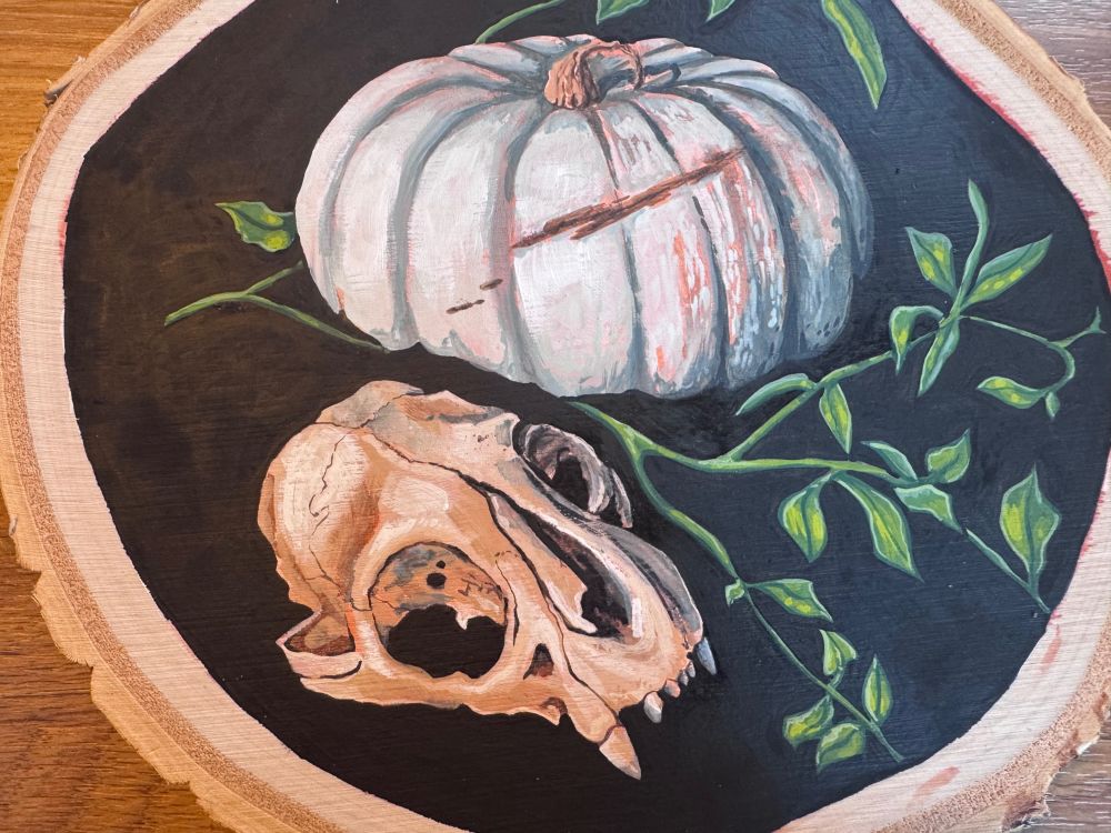 A circular Gouache painting on wooden birch slice. Depicting a small animal skull, green foliage and a white pumpkin on a black background. 

The paintings sits n a wooden countertop.
