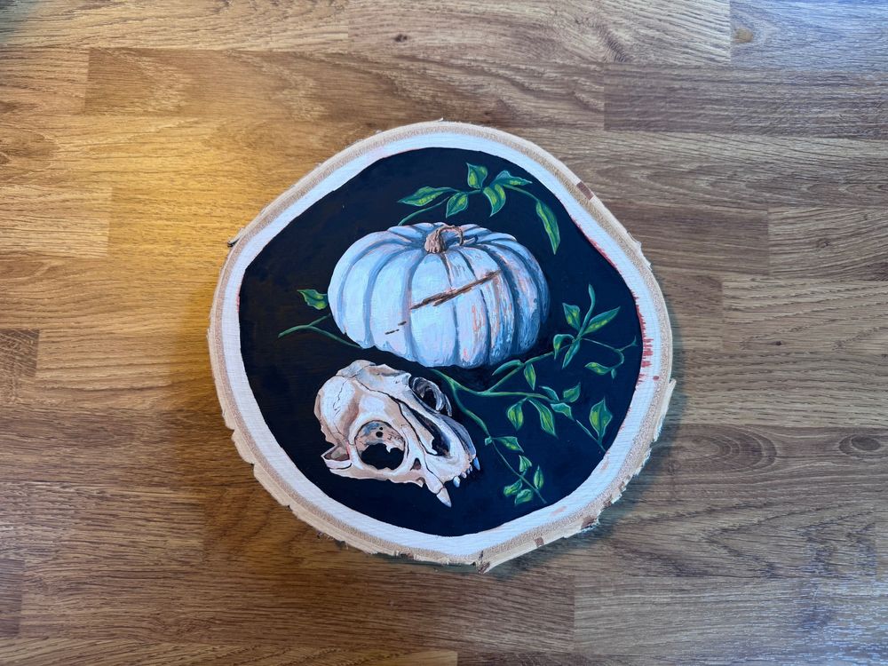 A circular Gouache painting on wooden birch slice. Depicting a small animal skull, green foliage and a white pumpkin on a black background. 

The paintings sits n a wooden countertop.
