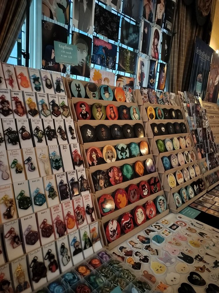 A different angle of the left and middle of the table where you can see keychains, coasters, badges, stickers and sticker sheets.