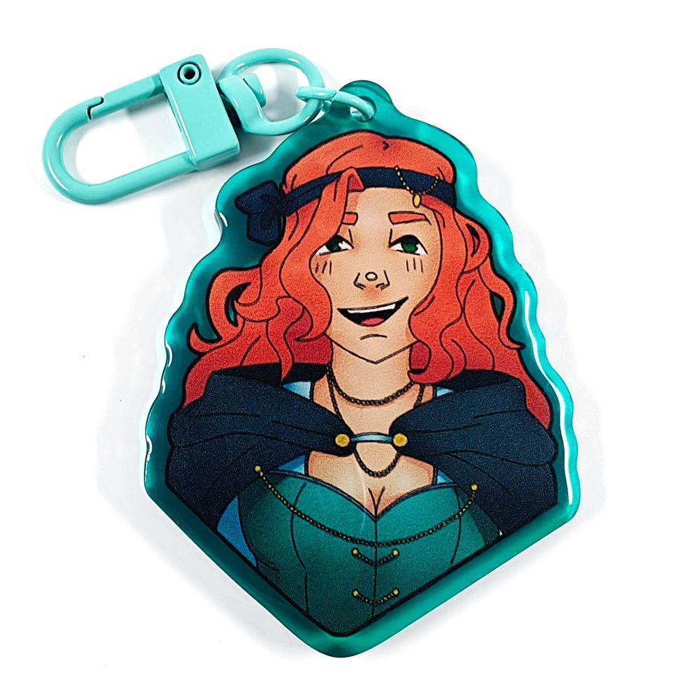 A picture of an acrylic keychain of GeminiTay with her pirate outfit. She has an open mouth smile. She is wearing a blue over the shoulders cape on top of a teal corset. She has a blue bandana in her hair, which is long, curly and orange. She is wearing different gold jewellery. The rim of the keychain is blue and it has a bright teal clasp.