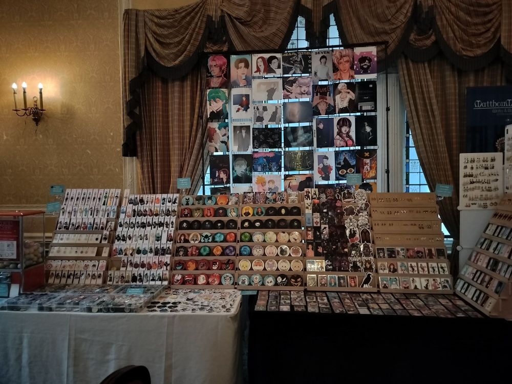 A picture of a double table set-up at Brighton Anime and Gaming Con. You can see handmade pendants, earrings, photocards on the right, sticker sheets, coasters and keychains in the middle and on the left. On the table in front of the keychains on the left there are six boxes full of badges. On the rest of the table space there are a lot of different stickers and some more photocards. Thee are a lot of art prints hung high behind the table. You can see merch of different fandoms like Hermitcraft, Hazbin Hotel, Ghibli, Don't Starve, QSMP, Vocaloid, League of Legends, Generation Loss, ATEEZ, Tower of God and more.