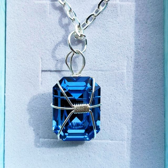A picture of a tall hexagonal blue crystal done with silver wire wrapping. There is a tight wrapping at the centre and three wires go out of it on each side, wrapping around the back of the crystal. At the top of it there is a loop attached to a silver ring, attached to a metal chain. This necklace is in a white jewellery box, with a teal border on each side.