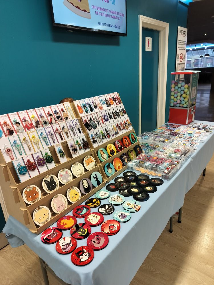A picture of a double table set-up at Teesside Comic Con. On the left you can see a lot of different keychains and coasters. On the middle of the table there are boxes full of badges. At the far end of the table, you can see a gacha machine, photocards and a lot of different stickers. You can see merch of a lot of different fandoms, like Stray Kids, ATEEZ, Hermitcraft, Hazbin Hotel, Don't Starve, Generation Loss, Ghibli, TXT, Loona, Xikers, NMIXX, G-Idle, Dreamcatcher, XDinary Heroes, BTS and more.