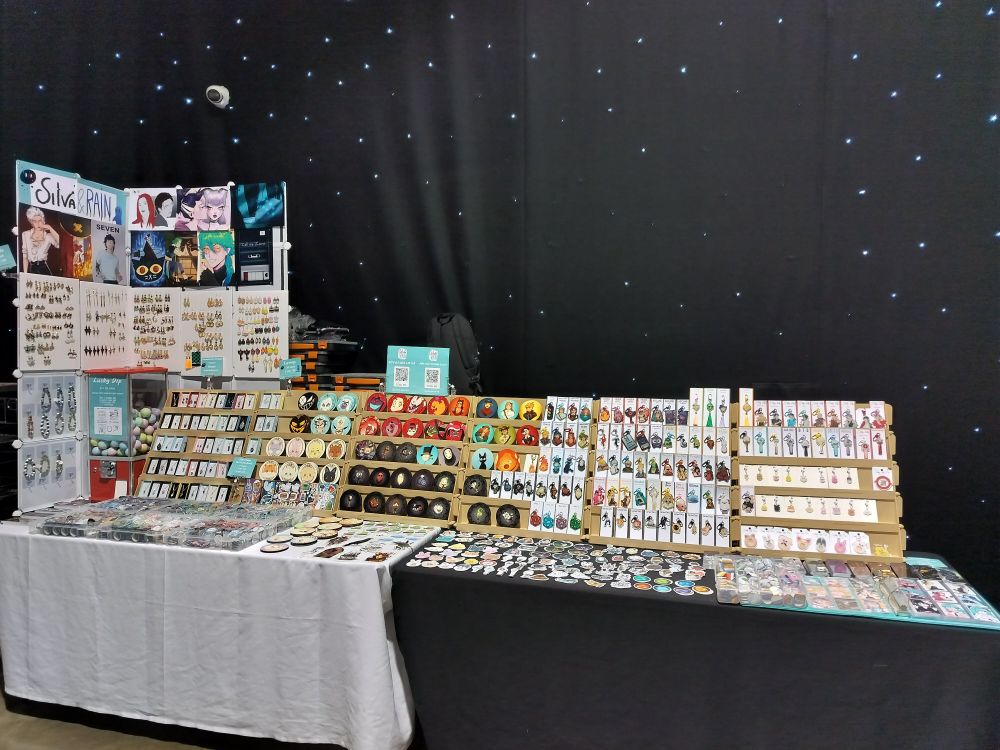 A picture of a double table set-up at Leeds Comic-Con. You can see handmade pendants, earrings, bracelets on the left, sticker sheets, coasters and keychains in the middle and on the right. On the table in front of the pendants on the left there are four boxes full of badges. On the rest of the table space there are a lot of different stickers and some more coasters, as well as a binder full of card size prints. You can see merch of different fandoms like Hermitcraft, Hazbin Hotel, Ghibli, Don't Starve, QSMP, Vocaloid, League of Legends, Generation Loss, ATEEZ, Tower of God and more.