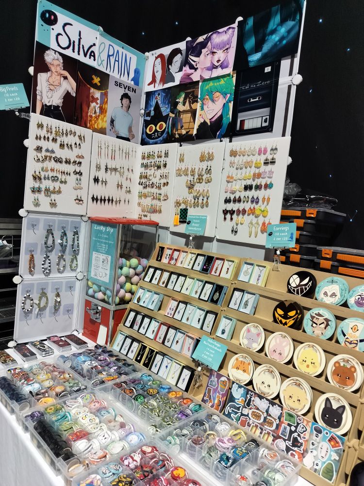A close-up of the left side of the tables where you can see prints, earrings, bracelets, necklaces, coasters, badges and sticker sheets.