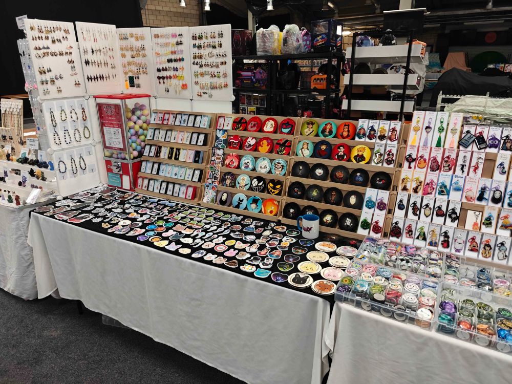 A picture of a double table set-up at Birmingham Anime and Gaming Con. You can see handmade pendants, earrings, bracelets on the left, sticker sheets, coasters and keychains in the middle and on the right. On the table in front of the keychains on the right there are four boxes full of badges. On the rest of the table space there are a lot of different stickers and some more coasters. You can see merch of different fandoms like Hermitcraft, Hazbin Hotel, Ghible, Don't Starve, QSMP, Vocaloid, League of Legends, Generation Loss, ATEEZ, Tower of God and more.