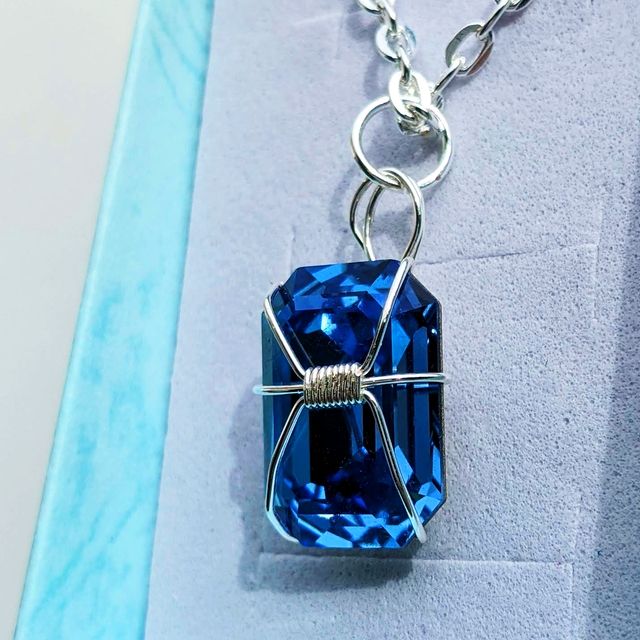 A picture of a tall hexagonal blue crystal done with silver wire wrapping, showb from a side angle. There is a tight wrapping at the centre and three wires go out of it on each side, wrapping around the back of the crystal. At the top of it there is a loop attached to a silver ring, attached to a metal chain. This necklace is in a white jewellery box, with a teal border on each side.