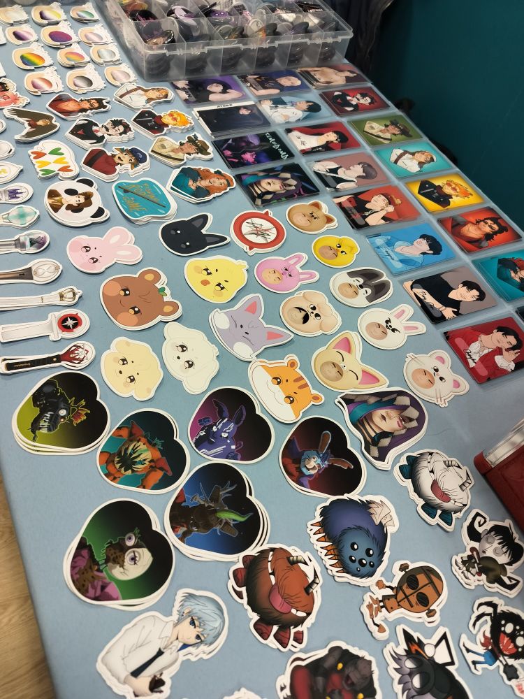 A close-up picture of the table at Teesside Comic-Con. You can see a lot of different stickers, including fandoms like Five Nights at Freddy's, ATEEZ, Stray Kids, Don't Starve, Hermitcraft and a lot of different K-Pop groups. You can also see some pride stickers, and some drawn photocards.