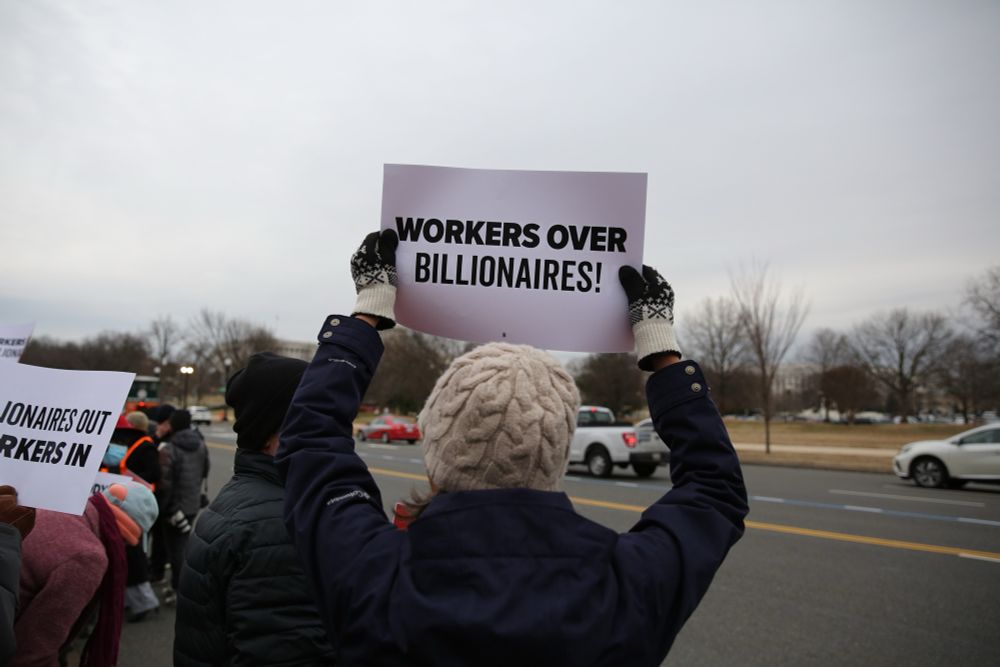 A photo of a protestor in front of the Department of Labor, holding a sign that says “workers over billionaires.”