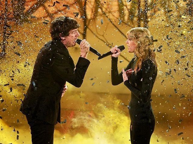 A picture of Taylor swift on the right and Gary lightbody on the left singing to each other, both wearing all black. The background is gold and there is gold confetti falling down beside them 