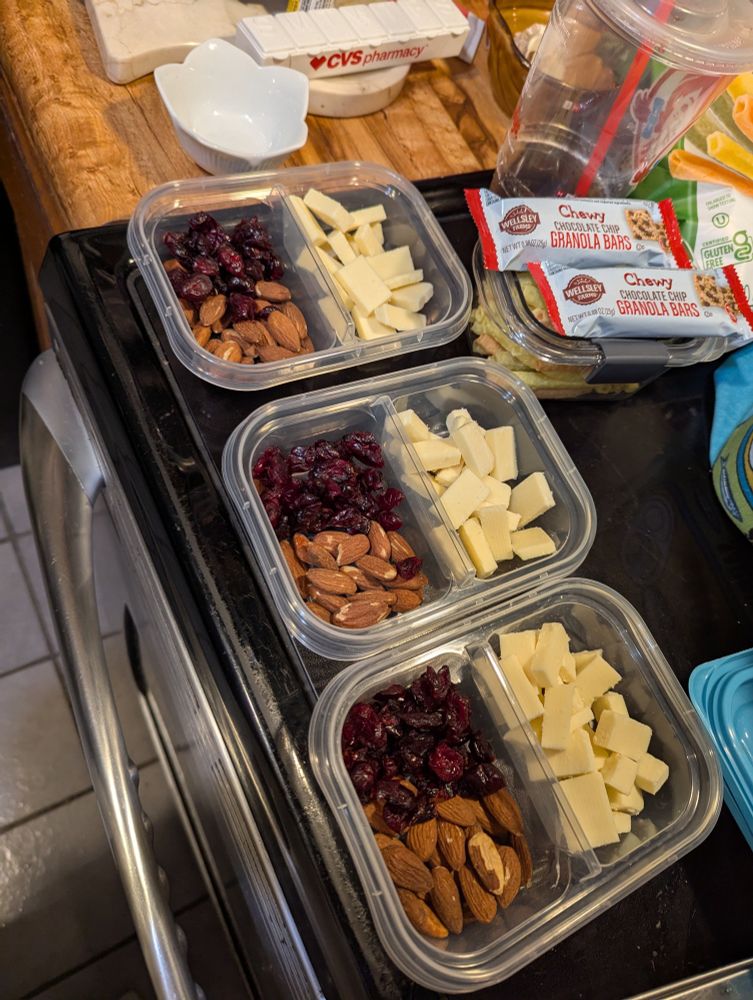 Three two-section snack tupperwares with cut up white cheddar, almonds, and craisins are lined up next to each other on a stovetop.