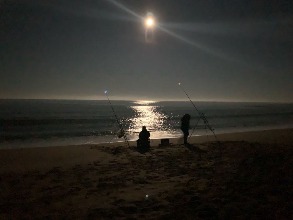 Two fishermen fishing with rods from Seaton Sluice beach by the light of the full moon.