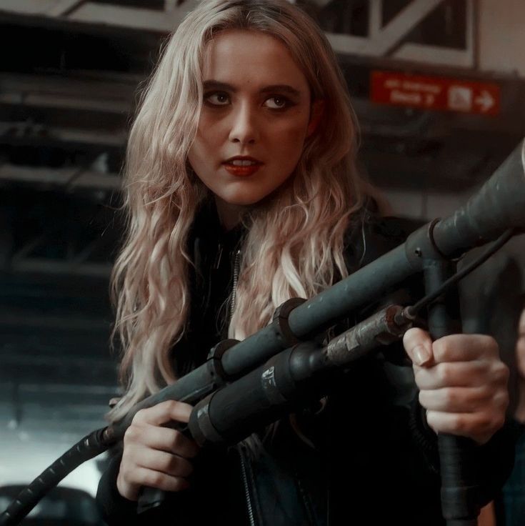 claire novak from supernatural holding a sick ass flamethrower. she's white, has long wavy blonde hair, and has black eyeliner and red lipstick. 