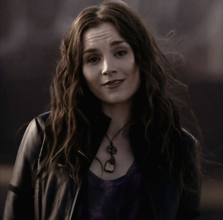 meg masters from season 5 of supernatural. she's white and has chest length, wavy dark brown hair, a purple shirt, a black leather jacket and a necklace. she's smirking.