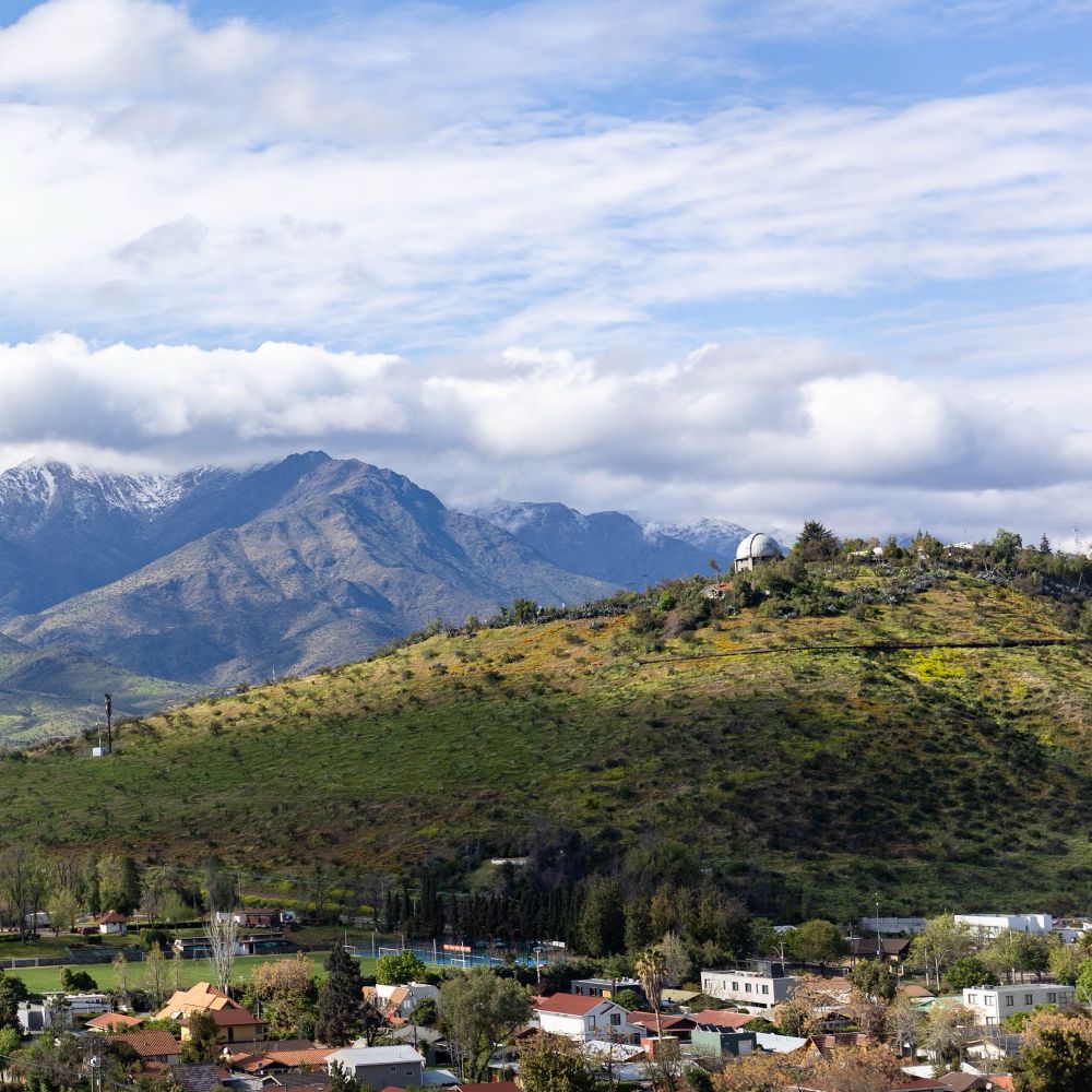 A green hillside rises above a neighborhood. A telescope dome is on top of the hill. In the background are tall mountains with snow and a blue sky with clouds. 