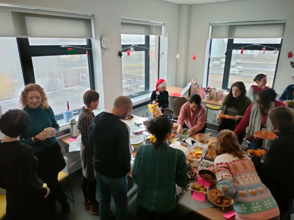 People chatting around a table laden with lots of different types of food. Pink pieces of paper show any allergies.