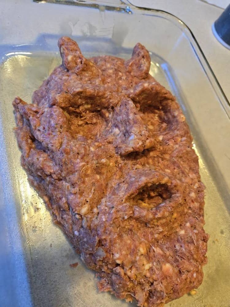 Raw meatloaf shaped like a head with horns, in a glass baking dish.