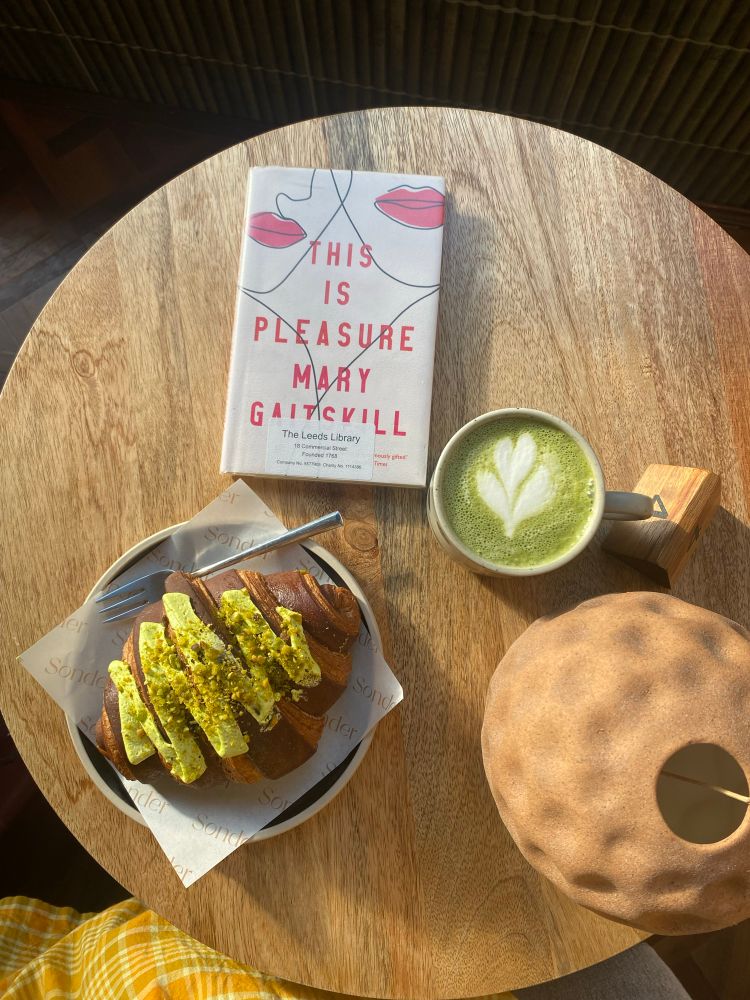 This image shows a cozy café setting with a round wooden table bathed in warm sunlight. On the table, there’s a copy of This Is Pleasure by Mary Gaitskill, its minimalist cream-colored cover featuring abstract line drawings of faces and lips in black and pink. The book is placed near the top left of the table.

Beside the book, to the right, is a cup of matcha latte in a ceramic mug, topped with delicate heart-shaped latte art. Below the book and latte, a golden-brown croissant is placed on a plate lined with branded parchment paper reading “Sonder.” The croissant is topped with vibrant green pistachio cream and crushed pistachios, giving it a textured appearance. A silver fork rests on the plate.

To the right of the latte, a textured, round, earth-toned object with a hollow opening sits on the table, possibly a decorative lamp or sculpture. In the bottom left corner of the image, part of a yellow gingham fabric—likely a skirt or dress—is visible, adding a soft contrast to the warm wooden tones of the table. The overall aesthetic is warm, inviting, and thoughtfully arranged.