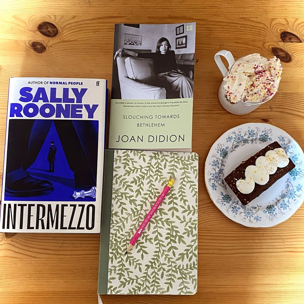 A cozy flat-lay on a wooden table featuring two books, a notebook, a mug of whipped cream-topped hot chocolate, and a slice of cake. The books are “Intermezzo” by Sally Rooney with a bold blue and black cover, and “Slouching Towards Bethlehem” by Joan Didion with a monochrome photograph of the author. A green floral-patterned notebook with a pink pencil lies beneath the books. To the right, there is a slice of chocolate cake with white frosting on a vintage floral-patterned plate, alongside a white mug with colorful sprinkles on the whipped cream. The setup exudes a warm and relaxing reading atmosphere.