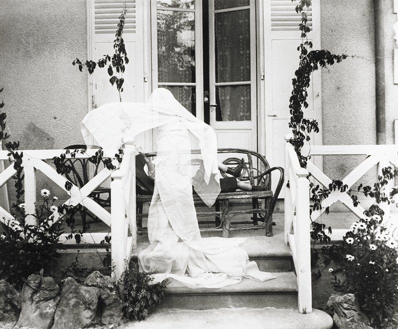 Black and white photograph of Lartigue's brother Maurice (know as Zissou) 'dressed as a ghost'. The image shows him outdoors, draped in white, ascending the steps of a veranda. A partly open door is ahead of him. 