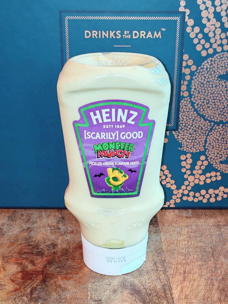 Photo: An squeezy bottle of Heinz Monster Munch Pickled Onion Flavour Mayo on a wooden worktop. The bottle has a purple label with the distinctive Monster Munch mascot and cartoon bats. Behind it sits a blue "Drinks by the Dram" gift box, because my kitchen has become a shrine to questionable flavour combinations. The mayo is pale yellow, which is either reassuring or deeply concerning depending on your relationship with pickled onions—and mine is clearly complicated.