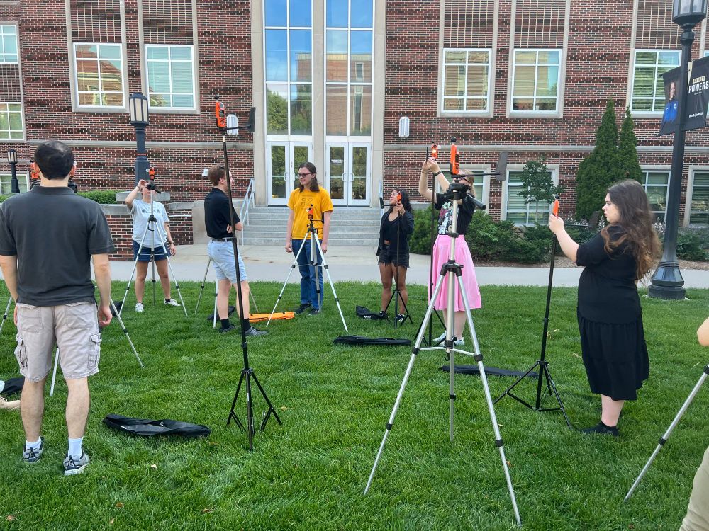 A group of people stand on grass, each manipulating a small weather instrument on a tripod.