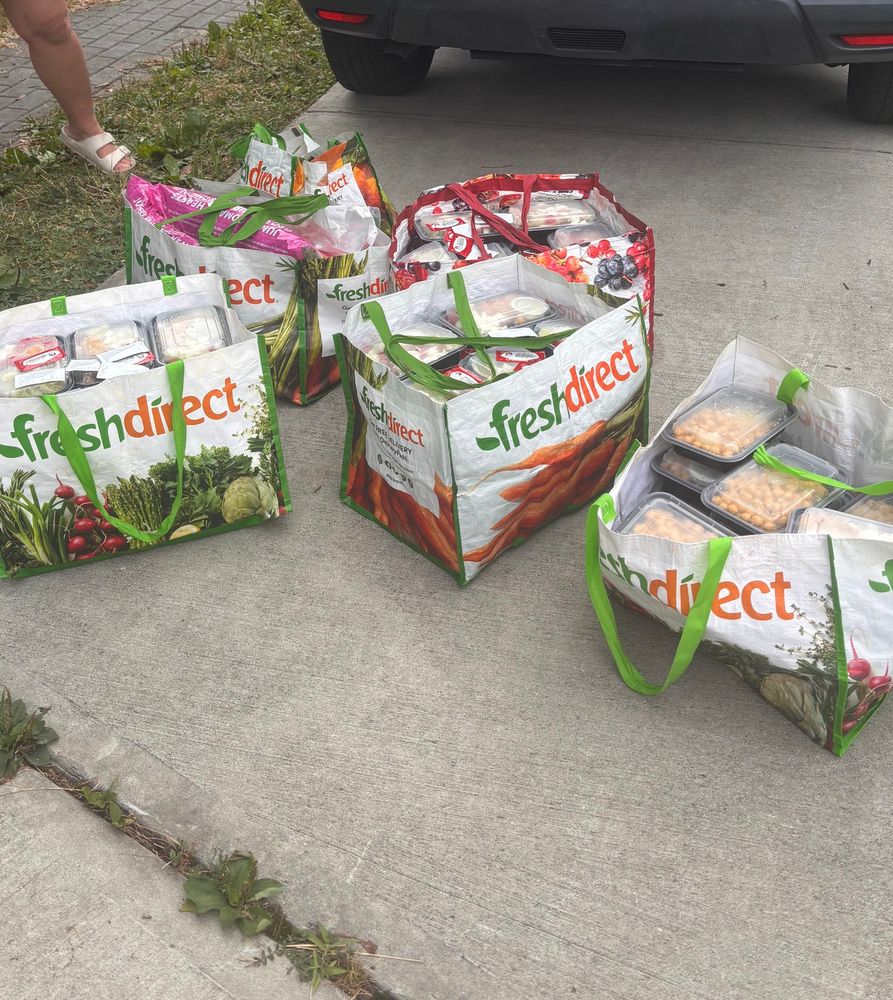 5 plastic tote bags filled with containers of hot meals on a piece of concrete behind a car. 