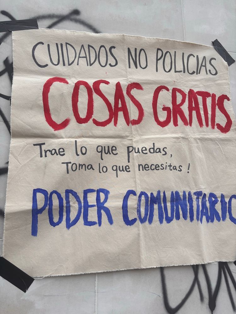 A white piece of canvas that says in red and blue paint “CUIDADOS NO POLICIAS COSAS GRATIS
Trae lo que puedas, Toma lo que necesitas !
PODER COMUNITARIO