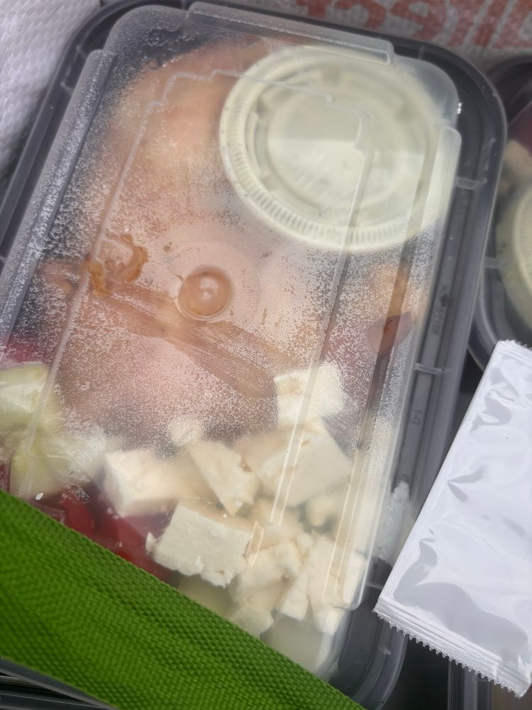 Plastic container steamed up. Cubes of feta, chicken and a container of greenish sauce visible. Hot sauce packet in foreground. 