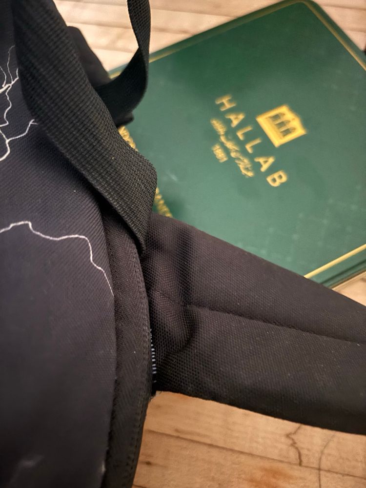A closeup of a black backpack strap on a wooden table with a visible repair. In the background,  a green & gold metal Hallab Lebanese sweets tin is blurry and one can presume it does not contain baklava 