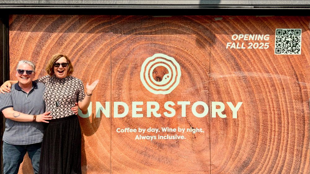 A man and a woman stand outside a storefront. The large sign in the window says “Understory. Coffee by day. Wine by night. Always inclusive. Opening fall 2025”